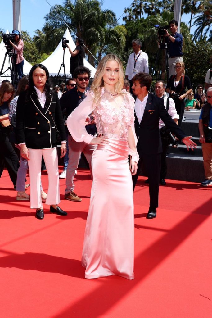 Blonde woman in a pink, floral-embellished gown posing on a red carpet as photographers and spectators surround her at a daytime event.