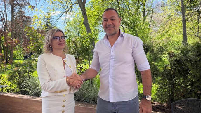 Man and woman shake hands outdoors in a sunny garden; she wears a white suit with gold buttons and glasses, he wears a light purple shirt and jeans.