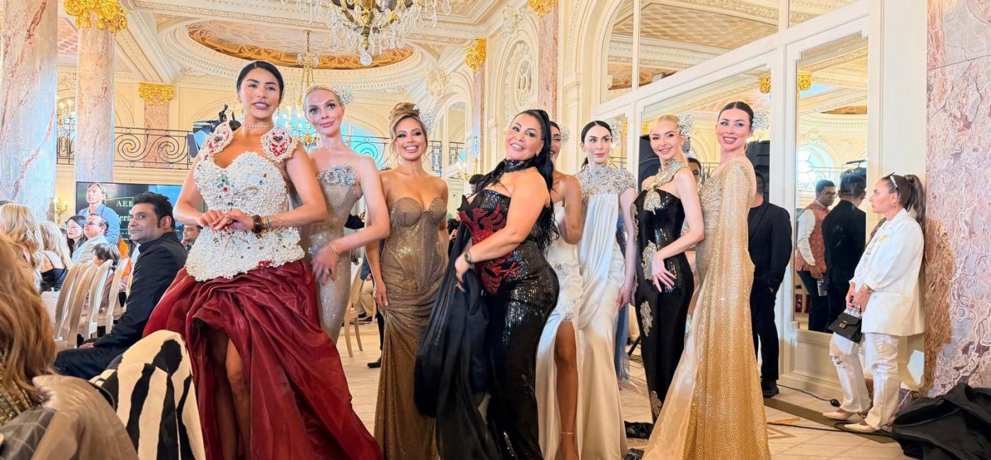 Group of models in glamorous gowns posing on a runway in an ornate ballroom with chandeliers and marble columns.