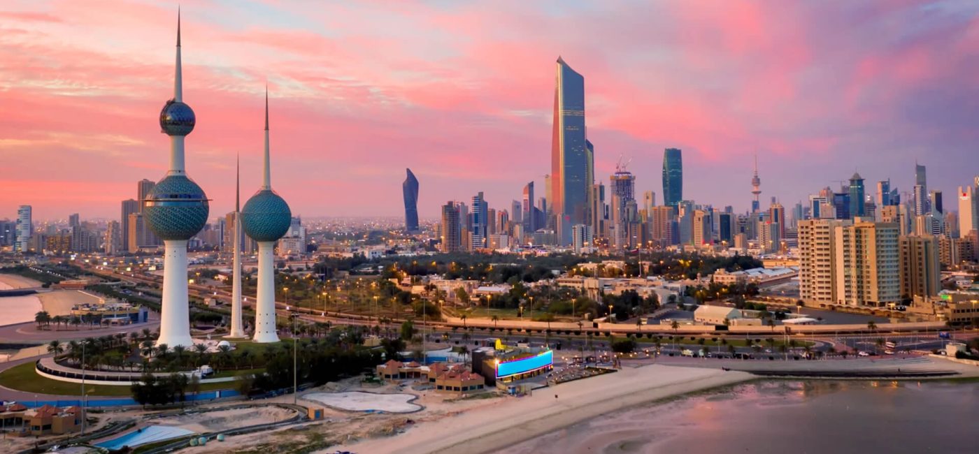 Kuwait City skyline at sunset with the iconic Kuwait Towers in the foreground and a mix of modern high-rise buildings behind them by the coastline