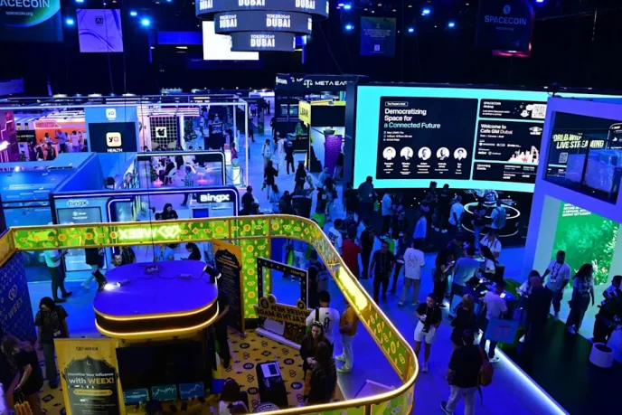 Wide view of a crowded tech expo floor with neon-lit booths, large digital screens, and attendees exploring colorful displays and kiosks.