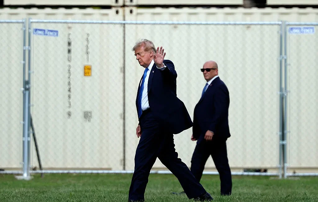 Man in a dark suit and blue tie waves while walking on grass, flanked by security in suits, with a chain-link fence behind.