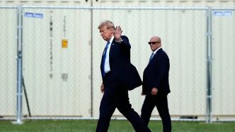 Man in a dark suit and blue tie waves while walking on grass, flanked by security in suits, with a chain-link fence behind.