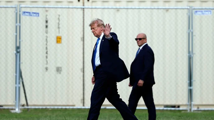 Man in a dark suit and blue tie waves while walking on grass, flanked by security in suits, with a chain-link fence behind.