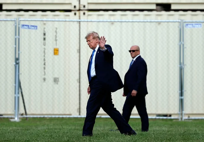 Man in a dark suit and blue tie waves while walking on grass, flanked by security in suits, with a chain-link fence behind.