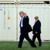 Man in a dark suit and blue tie waves while walking on grass, flanked by security in suits, with a chain-link fence behind.