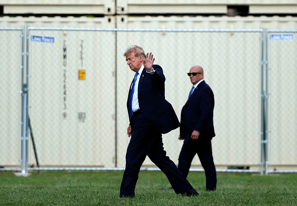 Man in a dark suit and blue tie waves while walking on grass, flanked by security in suits, with a chain-link fence behind.
