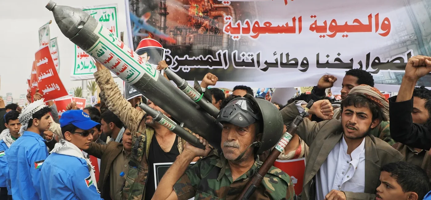 Crowd of protesters marching with a large rocket prop, raised fists, and a big Arabic banner in the background.