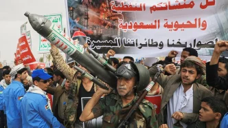 Crowd of protesters marching with a large rocket prop, raised fists, and a big Arabic banner in the background.