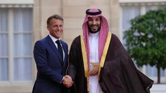 Two men in formal attire shake hands and smile outside a building; one in a navy suit, the other in traditional Arab dress with a gold-trimmed cloak and red-checked headscarf.