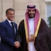 Two men in formal attire shake hands and smile outside a building; one in a navy suit, the other in traditional Arab dress with a gold-trimmed cloak and red-checked headscarf.