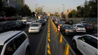 Heavy traffic on a city highway with a central yellow barrier, cars in both directions, and construction cranes in the background.
