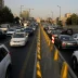 Heavy traffic on a city highway with a central yellow barrier, cars in both directions, and construction cranes in the background.