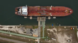 Aerial view of a red cargo vessel moored at a dock with green water and complex piping on deck.