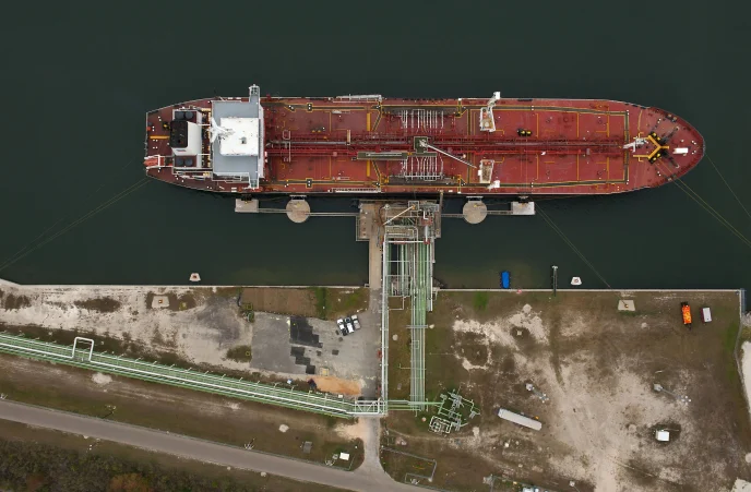 Aerial view of a red cargo vessel moored at a dock with green water and complex piping on deck.