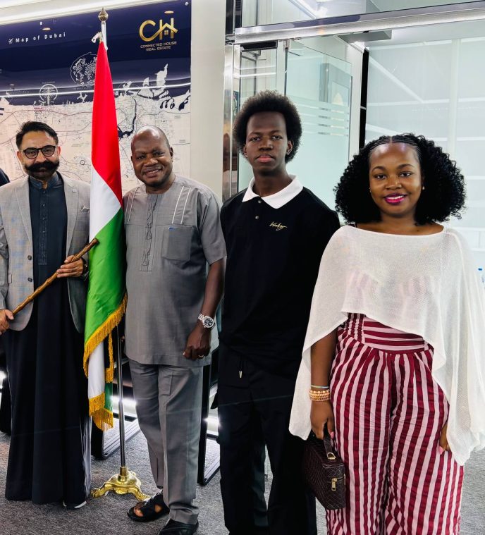 Four people pose indoors with a Nigerian flag between them: two men in traditional and casual attire, a young man in a black polo, and a woman in white poncho and red-striped pants.