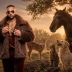 A man in a fur coat and sunglasses stands with a dog, a cat, and a horse in a sunset pasture.