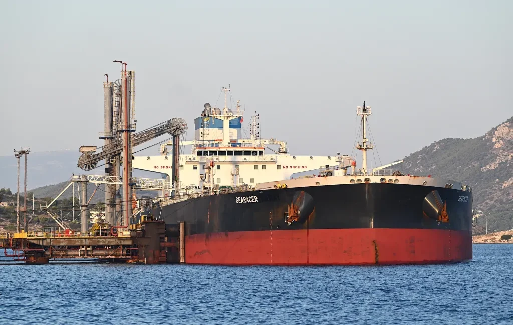 Large bulk carrier Searacer docked at a port with industrial loading equipment and hills in the background