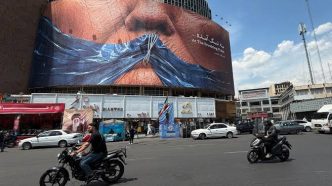 Busy urban street in front of a building under construction with a large curved billboard showing a close-up of a face; cars, motorcycles, and pedestrians move along the road.