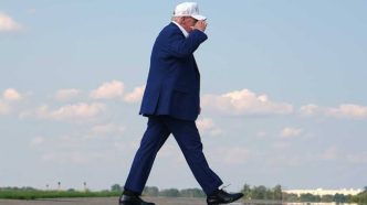 Man in a navy suit and white cap walking across an open airstrip under a blue sky, clouds in the background.