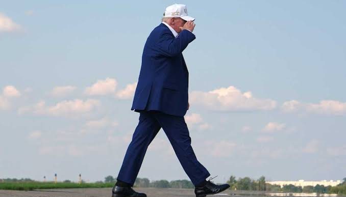 Man in a navy suit and white cap walking across an open airstrip under a blue sky, clouds in the background.