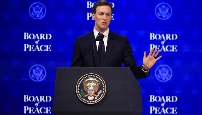Man in a dark suit speaks from a podium with the presidential seal at a Board of Peace event, gesturing with one hand raised.
