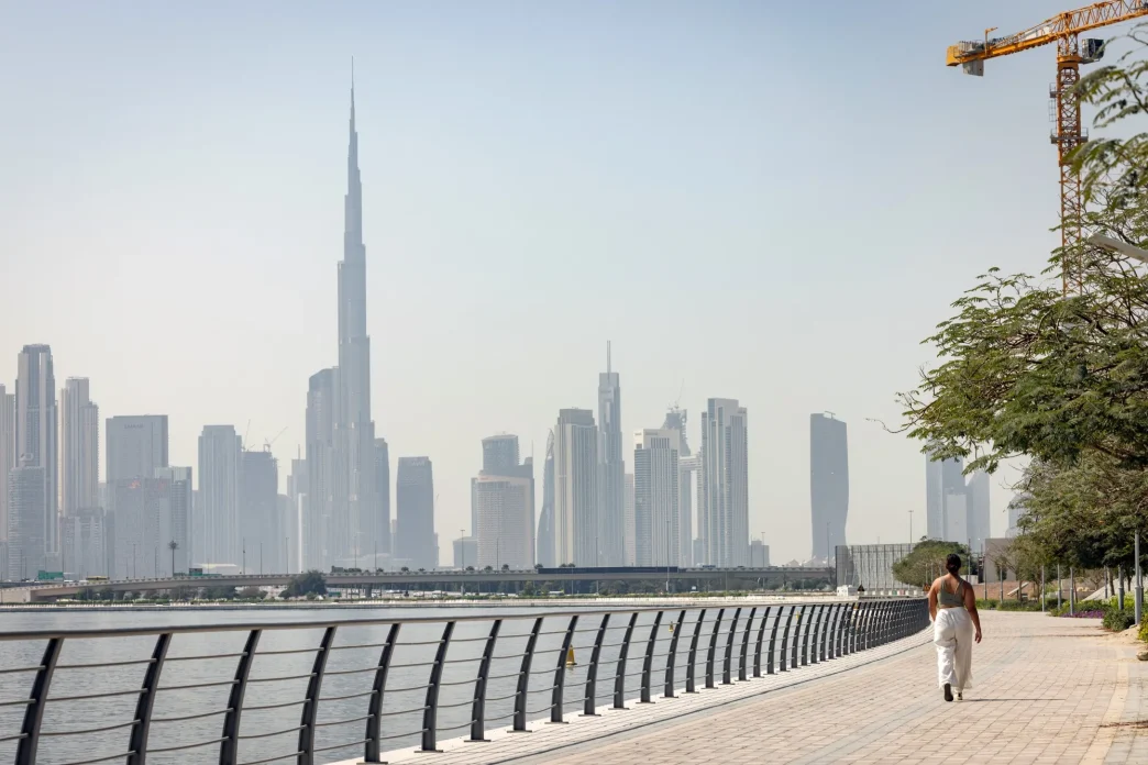 A person walking along a waterfront promenade with the Dubai skyline behind