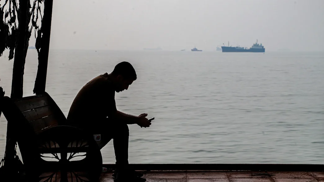A man sits by the Persian Gulf as cargo ships pass in the distance