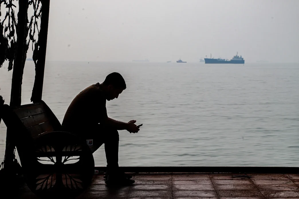 A man sits by the Persian Gulf as cargo ships pass in the distance