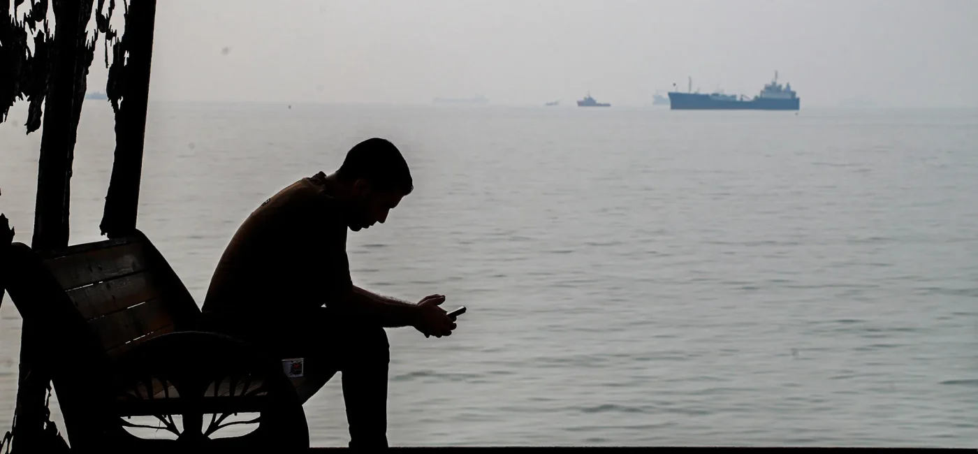 A man sits by the Persian Gulf as cargo ships pass in the distance