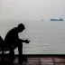 A man sits by the Persian Gulf as cargo ships pass in the distance